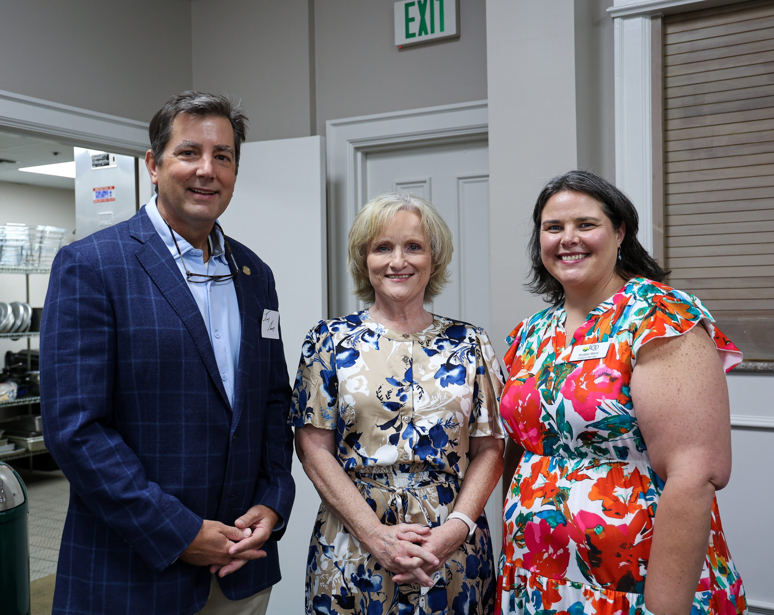 Three people pose together in what appears to be an office or institutional building, with an exit sign visible in the background. A man in a navy blazer and light blue shirt stands on the left, a woman with short blonde hair wearing a blue and white floral dress stands in the center, and a woman with dark hair wearing a bright multicolored floral dress stands on the right. All three are smiling at the camera.