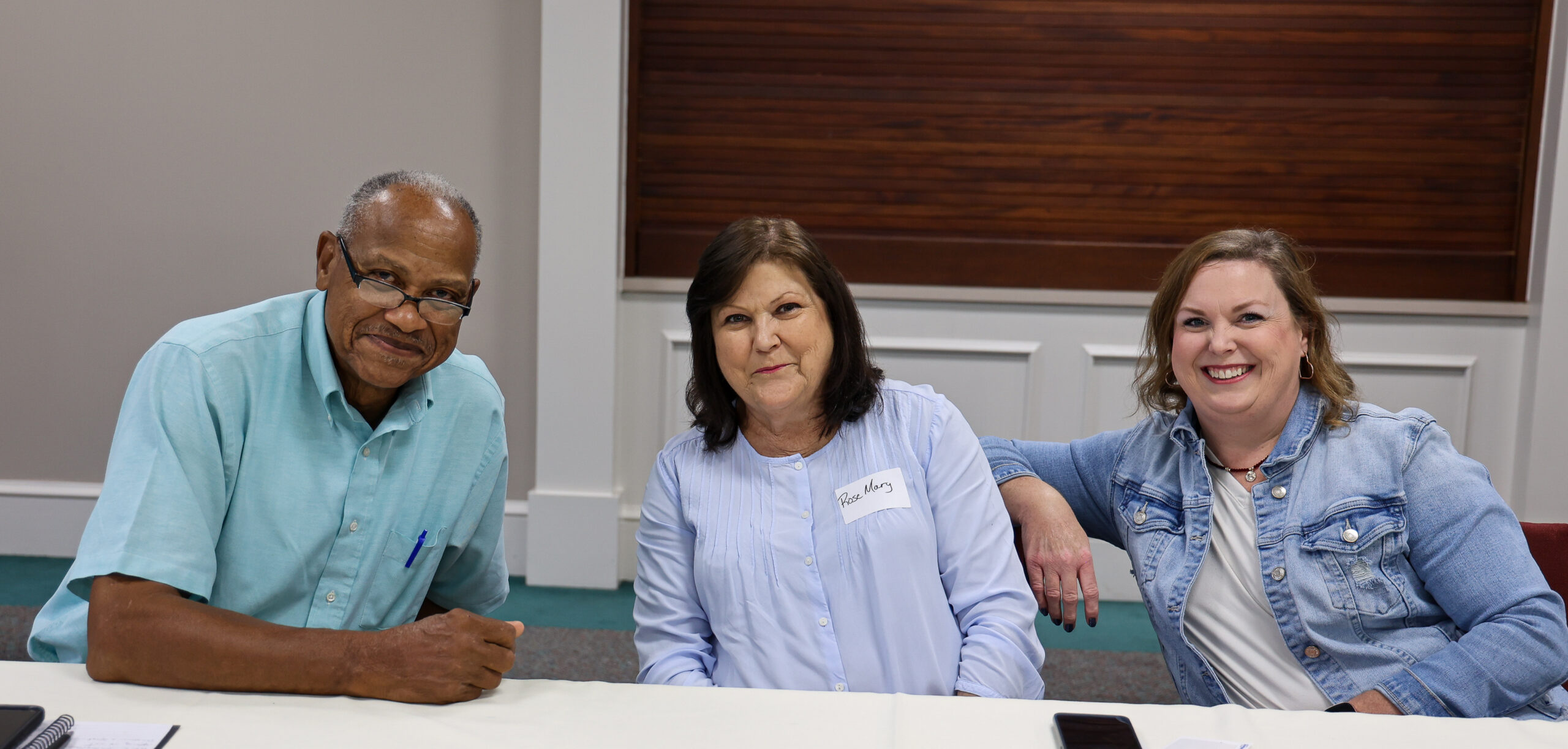Three people seated at a white table in what appears to be a conference room. A Black man in glasses and a light blue shirt sits on the left, a woman with dark hair in a light blue blouse sits in the center wearing a name tag, and a woman in a denim jacket sits on the right with her arm propped against the center person.