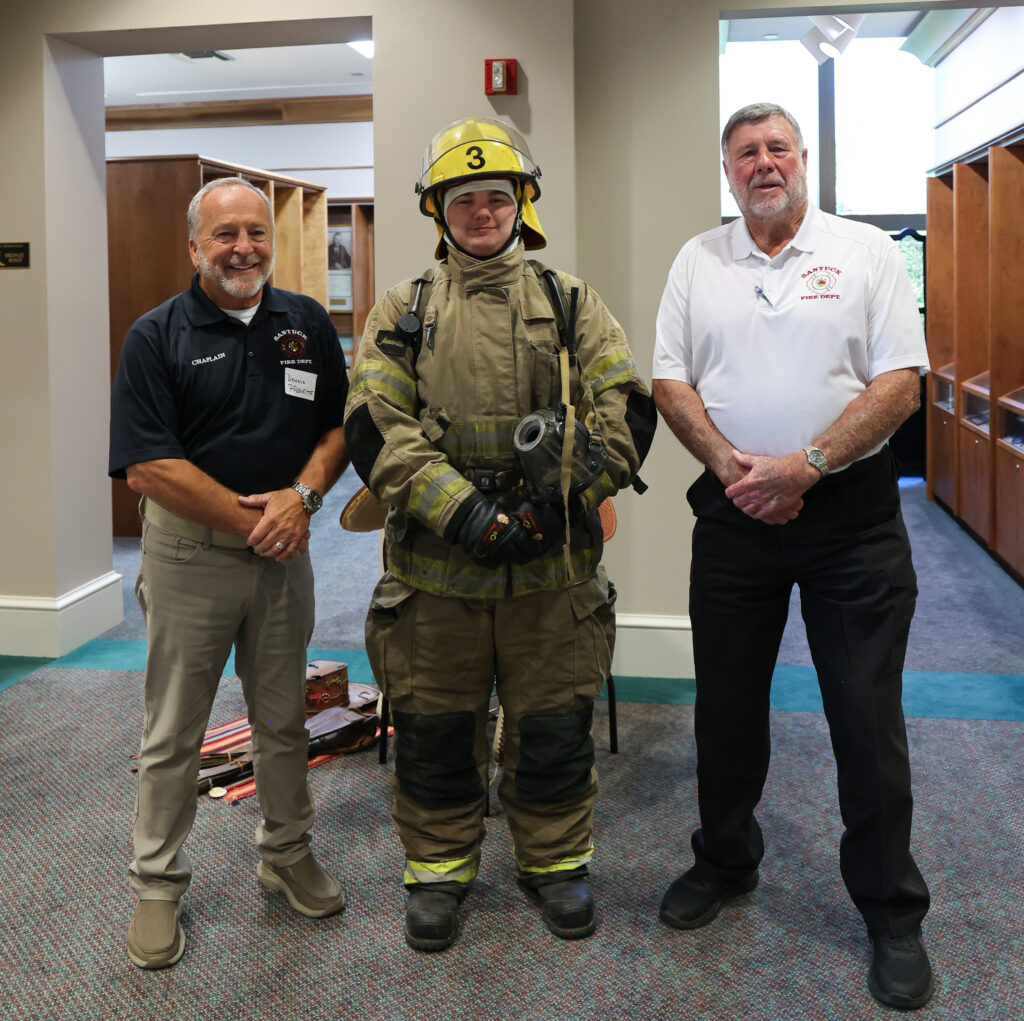 Three men standing in an office hallway. On the left, a man with a gray beard wearing a dark polo shirt and khaki pants. In the center, a firefighter in full gear including a yellow helmet marked "3," tan and yellow reflective turnout coat and pants, and breathing apparatus. On the right, an older man with a white beard wearing a white polo shirt with fire department logo and dark pants. The setting appears to be inside a government or municipal building.