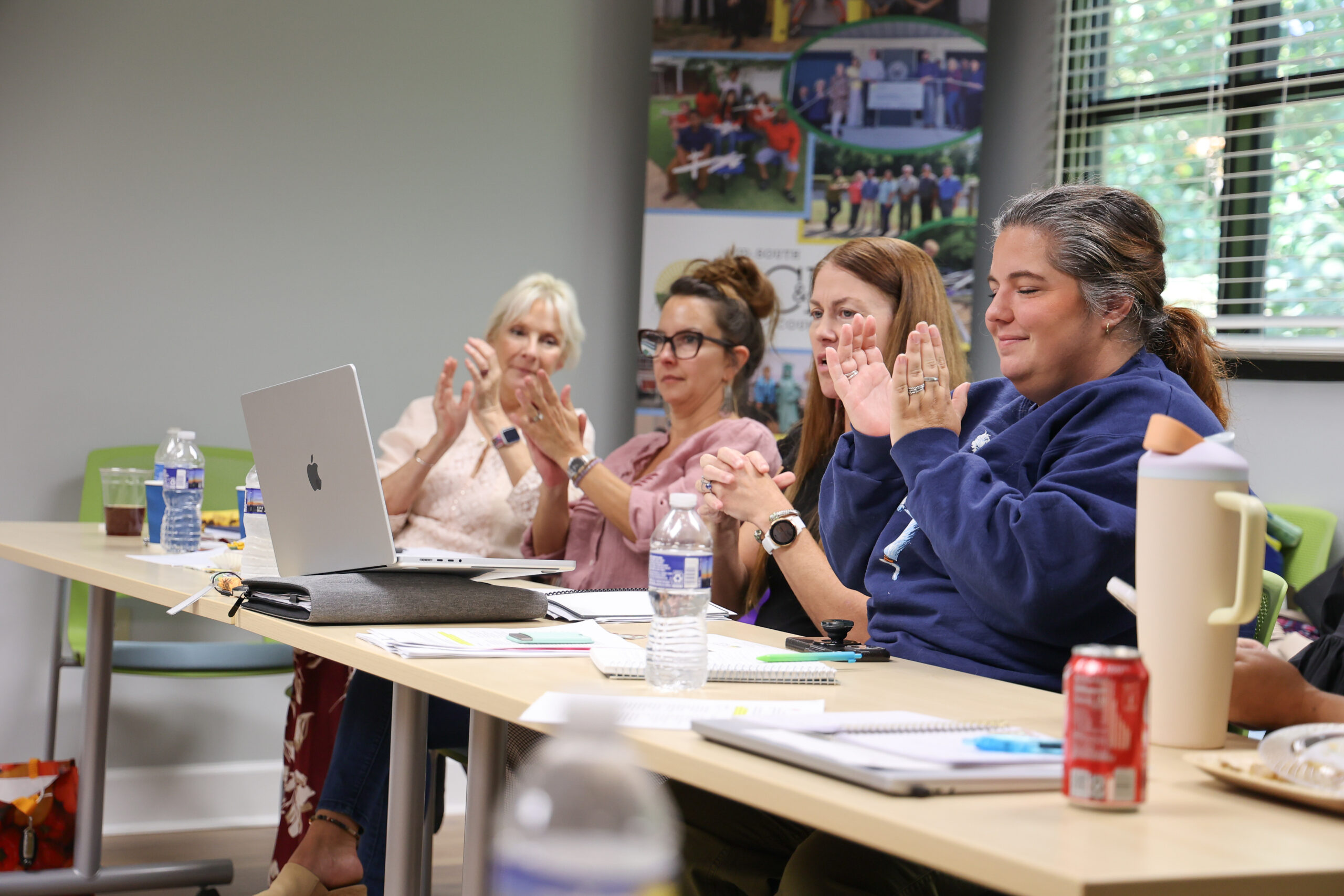 Four women seated at a table during what appears to be a meeting, with their hands raised as if clapping. From left to right: a blonde woman in a light top, a woman with glasses in a pink top, a woman with long hair, and a woman in a navy blue hoodie. A laptop, water bottles, and other meeting materials are visible on the table. A bulletin board with photos is visible on the wall behind them.