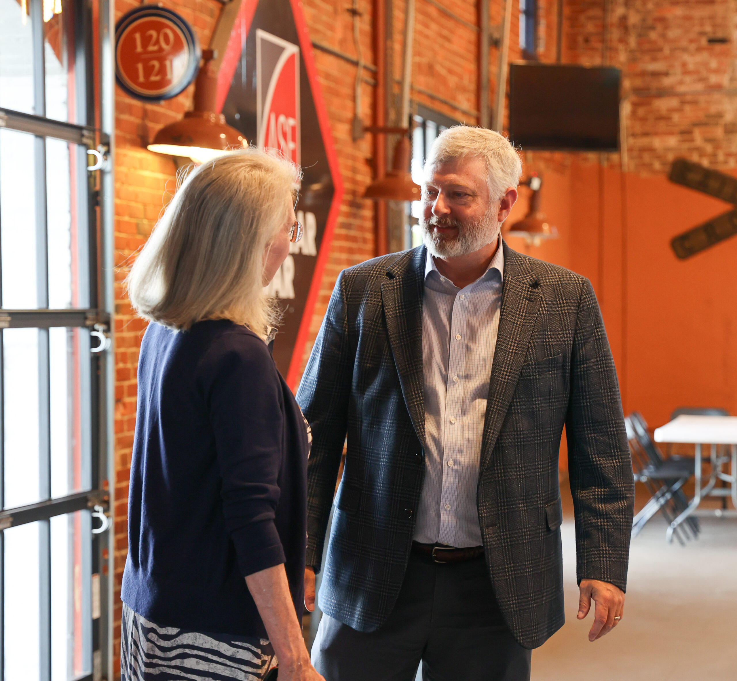 Man with white hair dressed in a gray suit coat and a pale button shit is speaking with a blonde woman with a navy sweater on. They appear to be in a sports bar with brick walls and sign decoration.