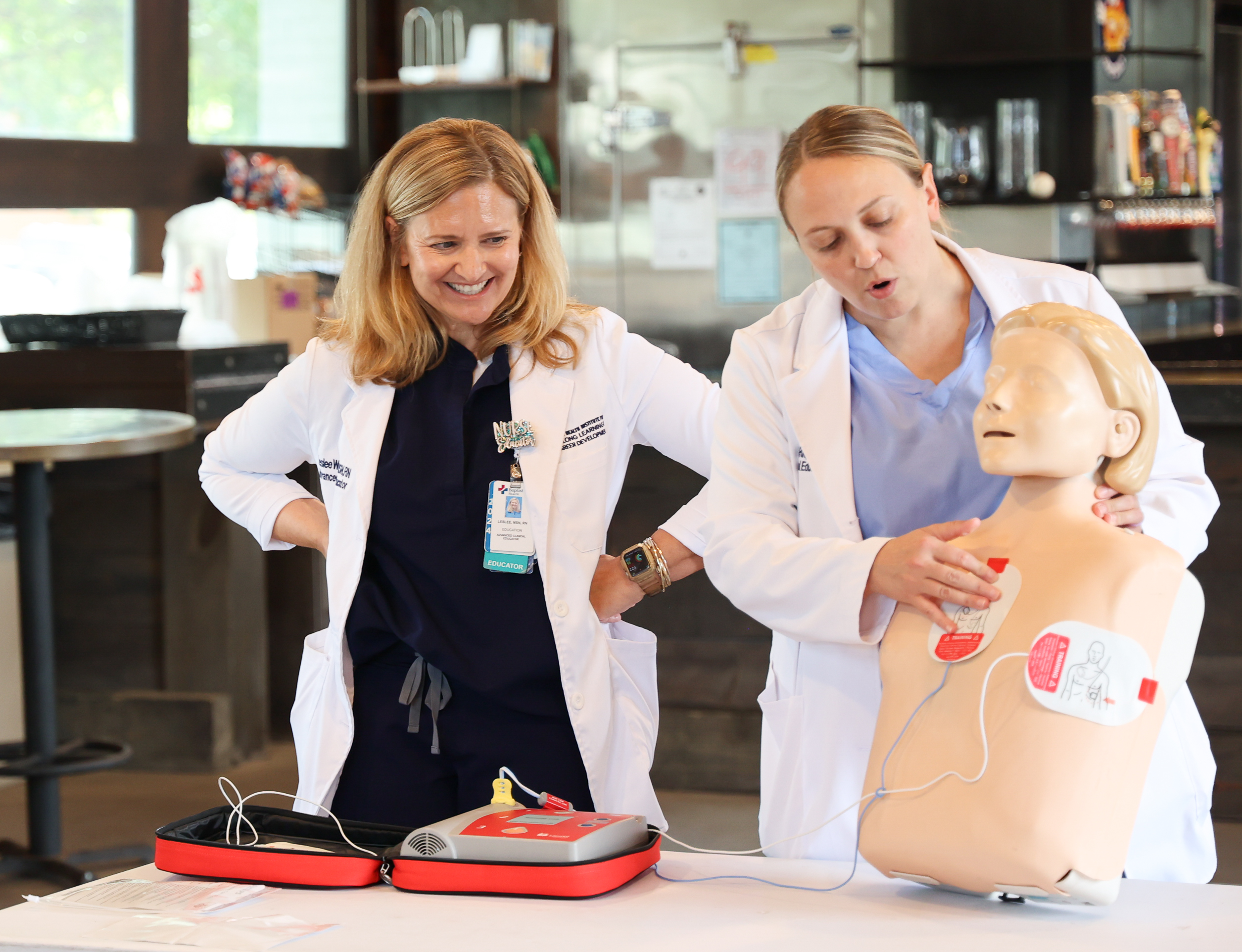 Two medical professionals in white lab coats and blue scrubs demonstrating AED training equipment. The woman on the left with blonde hair watches as the woman on the right positions a medical training mannequin with red electrode pads attached. A red automated external defibrillator (AED) device sits on the table in front of them. The setting appears to be a bar with a kitchen visible in the background.