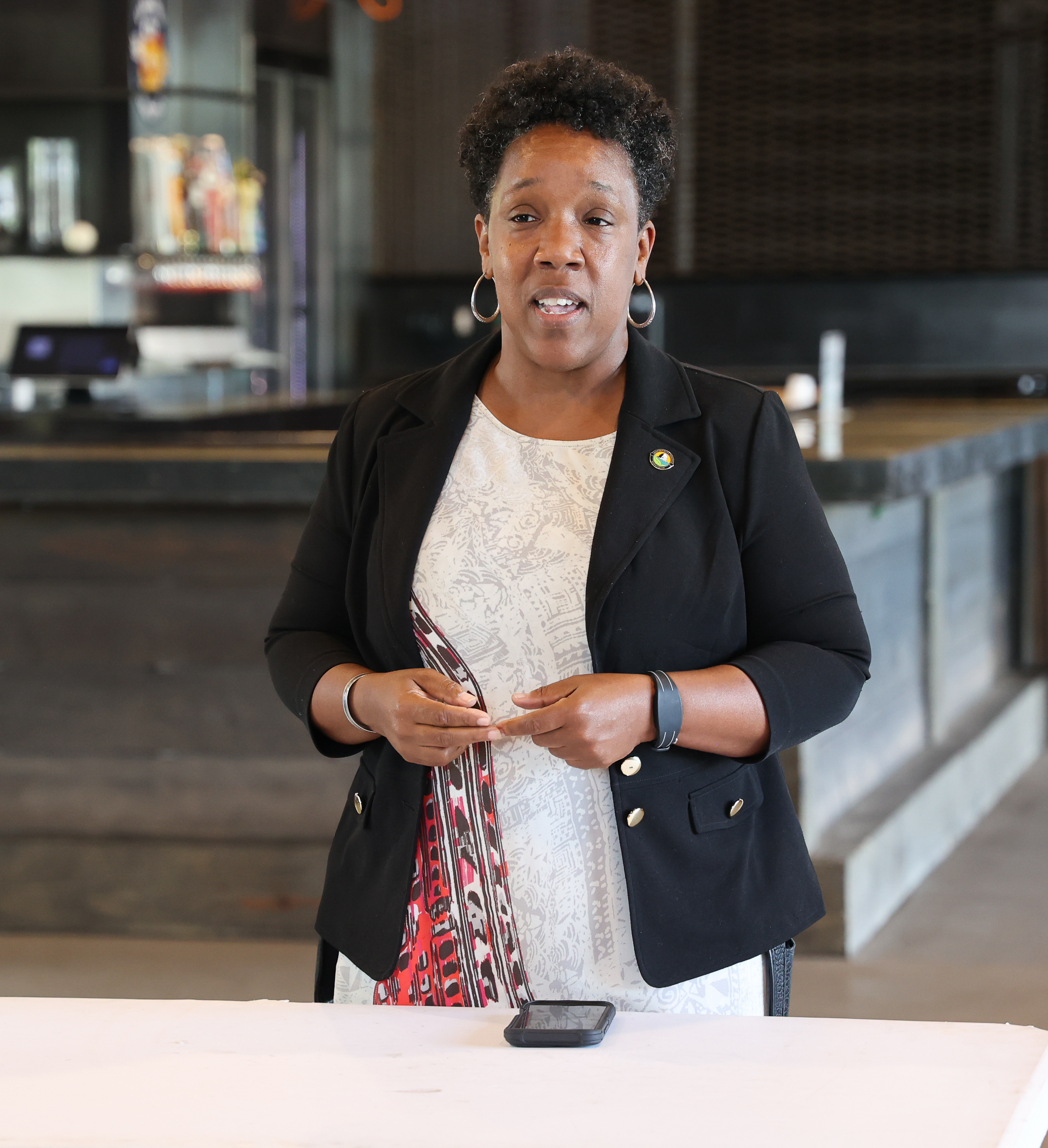 A woman speaking while standing behind a table. She is wearing a black blazer over a white patterned dress with red and black accents. She has short curly hair, hoop earrings, and a smartwatch. Behind her is a modern, industrial-style interior with a bar counter and dim lighting.