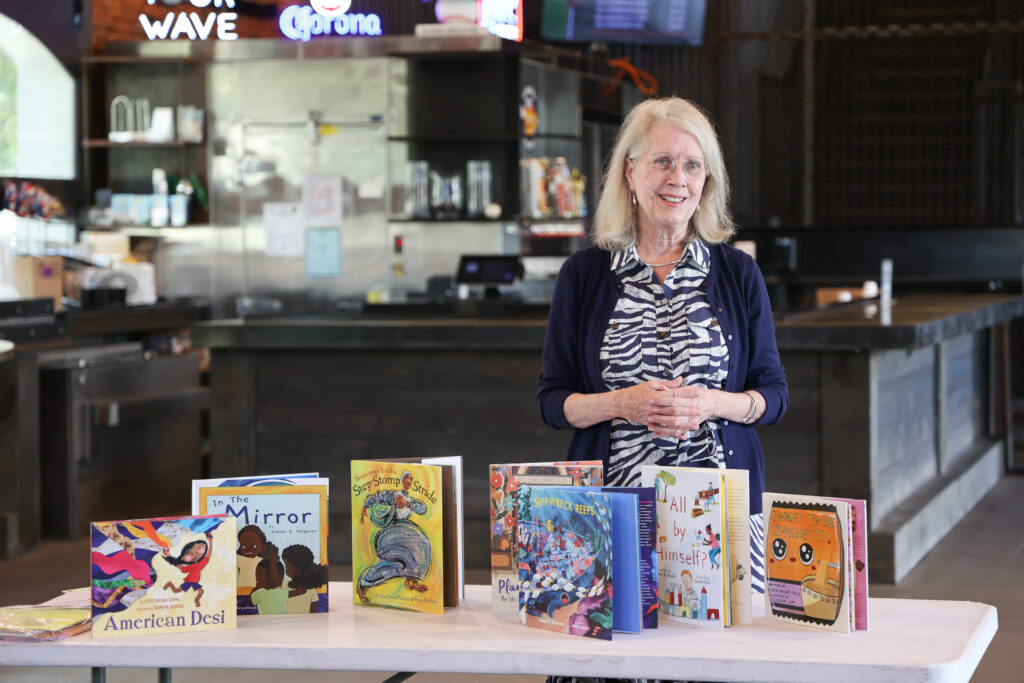 An older woman with long white hair stands smiling behind a table displaying colorful children's books. She is wearing a navy cardigan over a zebra-print blouse. The books include titles like American Desi, In the Mirror, Step Stomp Stride, and I Want to Be Spaghetti!. The background features a bar area with stainless steel appliances, taps, and neon beer signs.