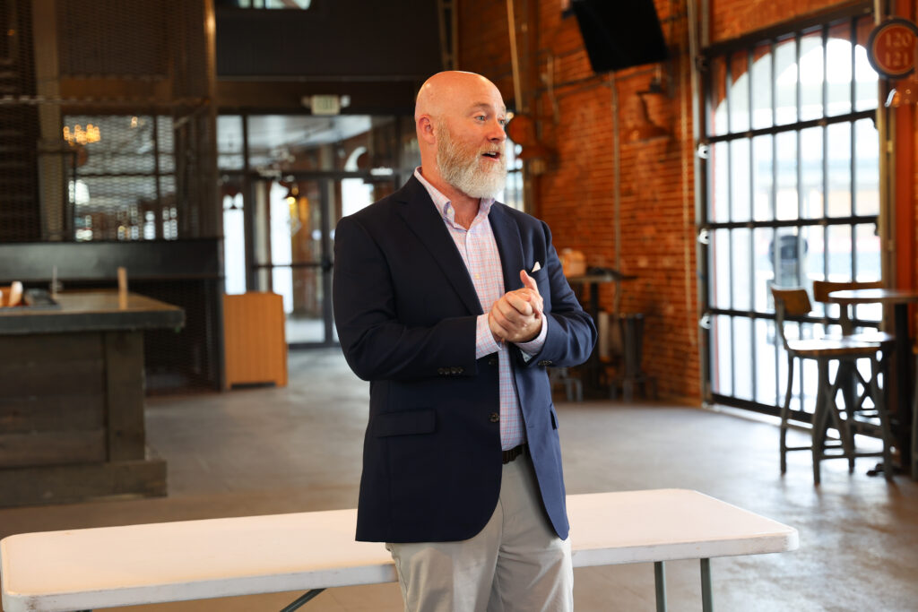 A man with a bald head and white beard wearing a navy blazer over a light checkered shirt stands speaking in an industrial-style space with exposed brick walls. He appears to be giving a presentation or talk.